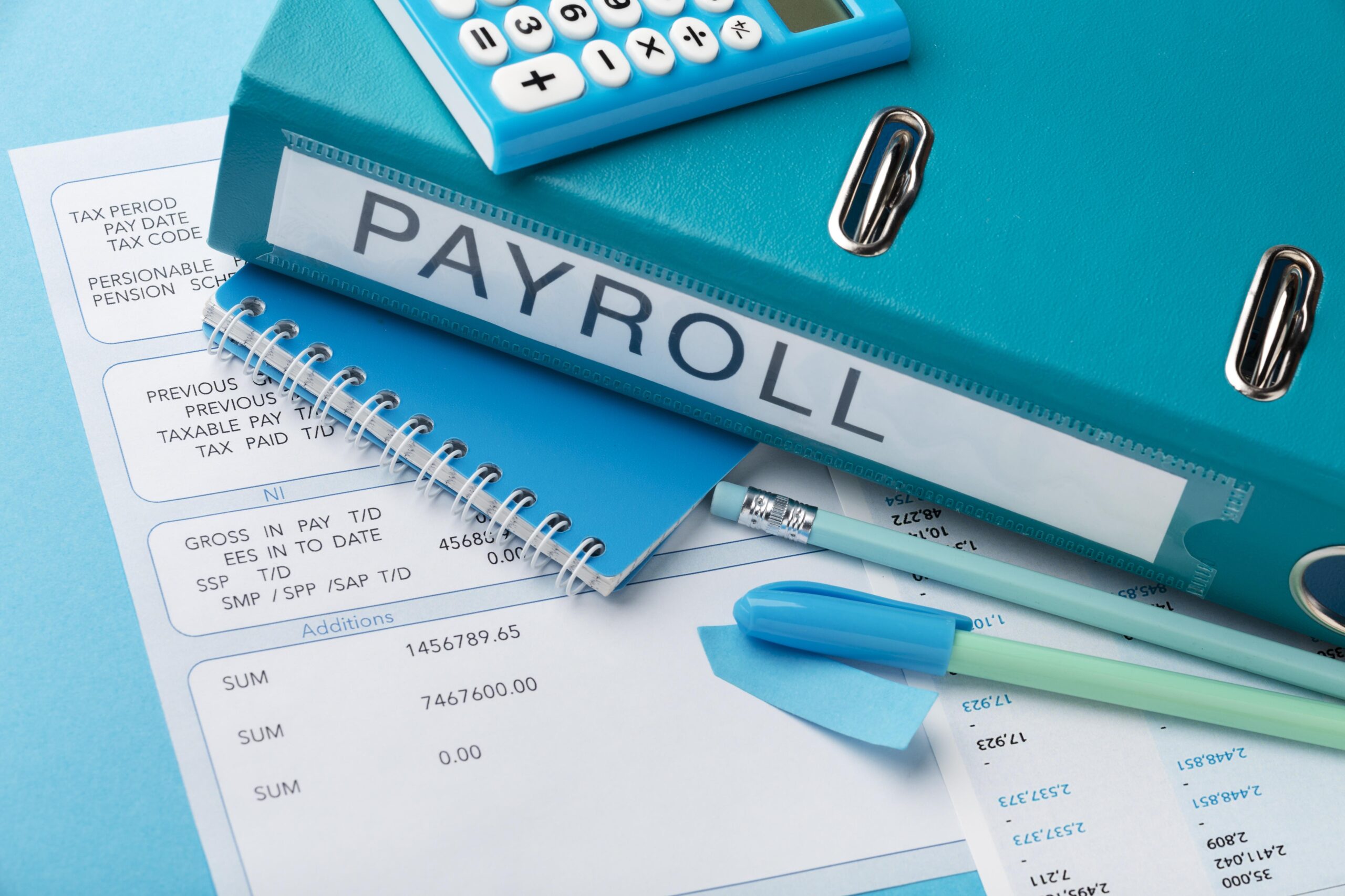 Close-up of payroll documents, a blue binder labeled 'PAYROLL', a calculator, a notebook, and various office supplies like pens, highlighters, and paper clips, all arranged on a blue background.